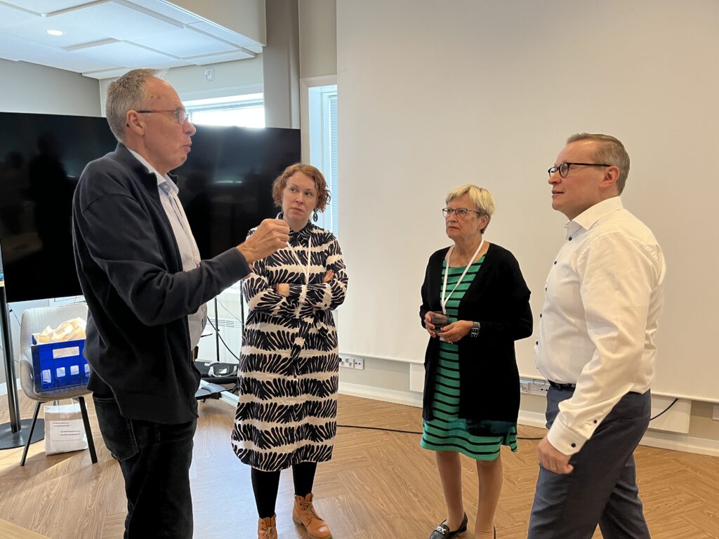 Fyra vuxna står i ett rum och samtalar. En man gestikulerar med handen medan de andra lyssnar. De är klädda i avslappnad kontorsklädsel. I bakgrunden syns kontorsutrustning och en stor skärm.