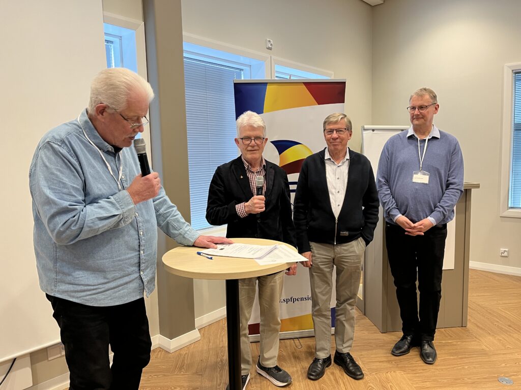 Fyra äldre män står inomhus. En man vid ett runt bord läser upp från ett papper i en mikrofon, medan de tre andra männen står tillsammans och vänder sig mot kameran. I bakgrunden syns en banderoll och fönster.