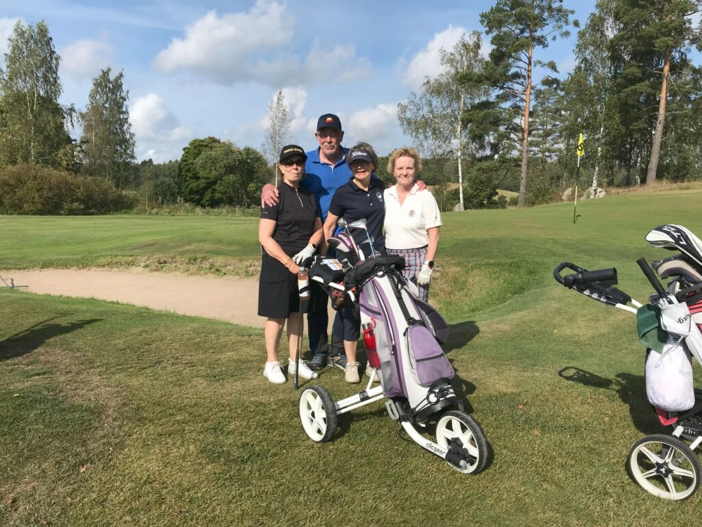 Fyra personer står på en golfbana nära en sandbunker och poserar och ler mot kameran. I förgrunden står två golfvagnar med väskor. I bakgrunden syns träd och en flaggstång.