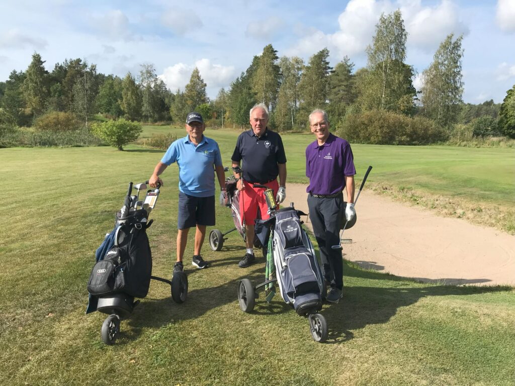 Tre män står på en golfbana bredvid sina golfväskor och vagnar. De är klädda i golfkläder och ler mot kameran, med träd och en sandbunker i bakgrunden.