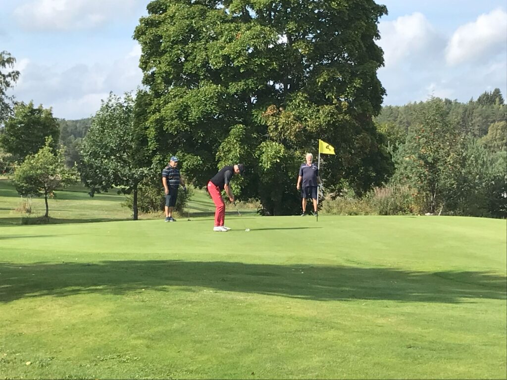 Tre personer på en golfbana; en puttar på greenen medan de två andra tittar på. Flaggstången står i hålet, och i bakgrunden syns träd och gräs under en delvis molnig himmel.