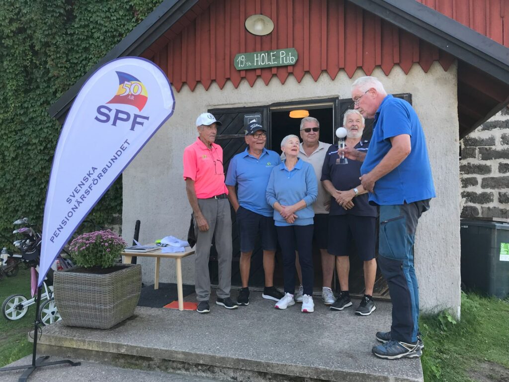 Sex äldre personer står utanför en byggnad med skylten ”H.O.L.E. Pub”. En man i blått visar upp något för gruppen. På en banderoll står det ”SPF Svenska Pensionärsförbundet 50 år”. I närheten står ett bord och golfutrustning.