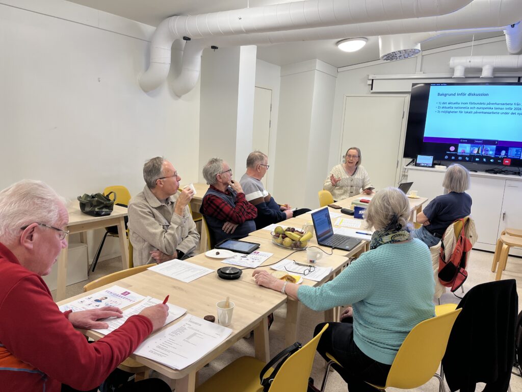 En grupp äldre vuxna sitter runt borden i ett klassrum och lyssnar på en kvinna som talar längst fram vid en skärm som visar en presentation. Papper, pennor och drycker finns på borden.
