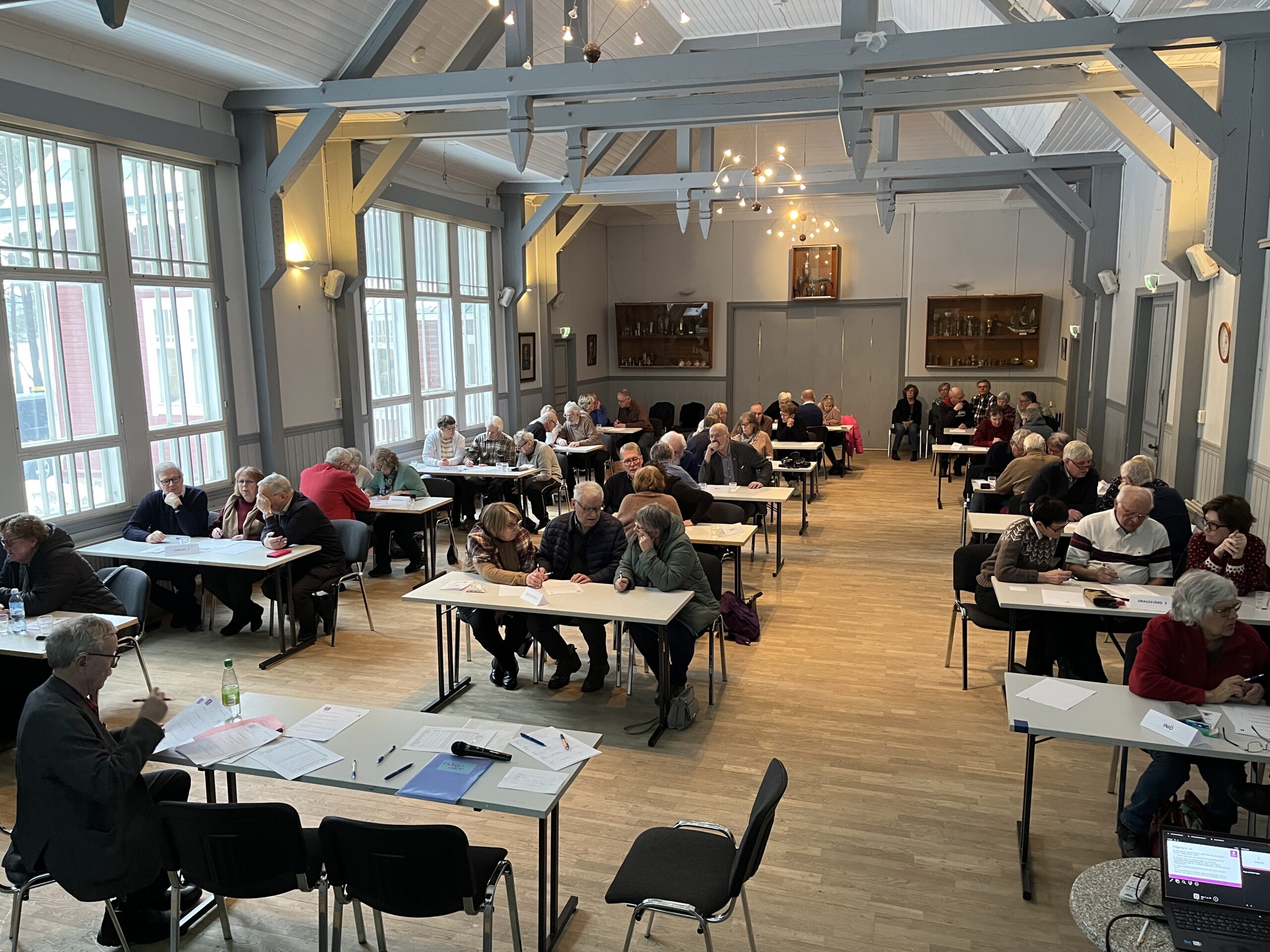 En grupp människor sitter vid bord i en rymlig, väl upplyst sal med stora fönster och högt i tak. De flesta deltagarna verkar vara upptagna med att prata eller titta på papper. En person står längst fram bredvid en bärbar dator.