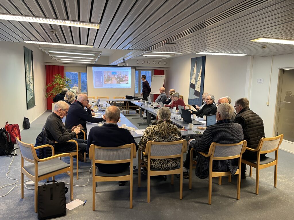 En grupp människor sitter runt ett rektangulärt bord i ett mötesrum, vända mot en föredragshållare som står bredvid en skärm där en presentation visas. På bordet ligger bärbara datorer, papper och kaffekoppar.