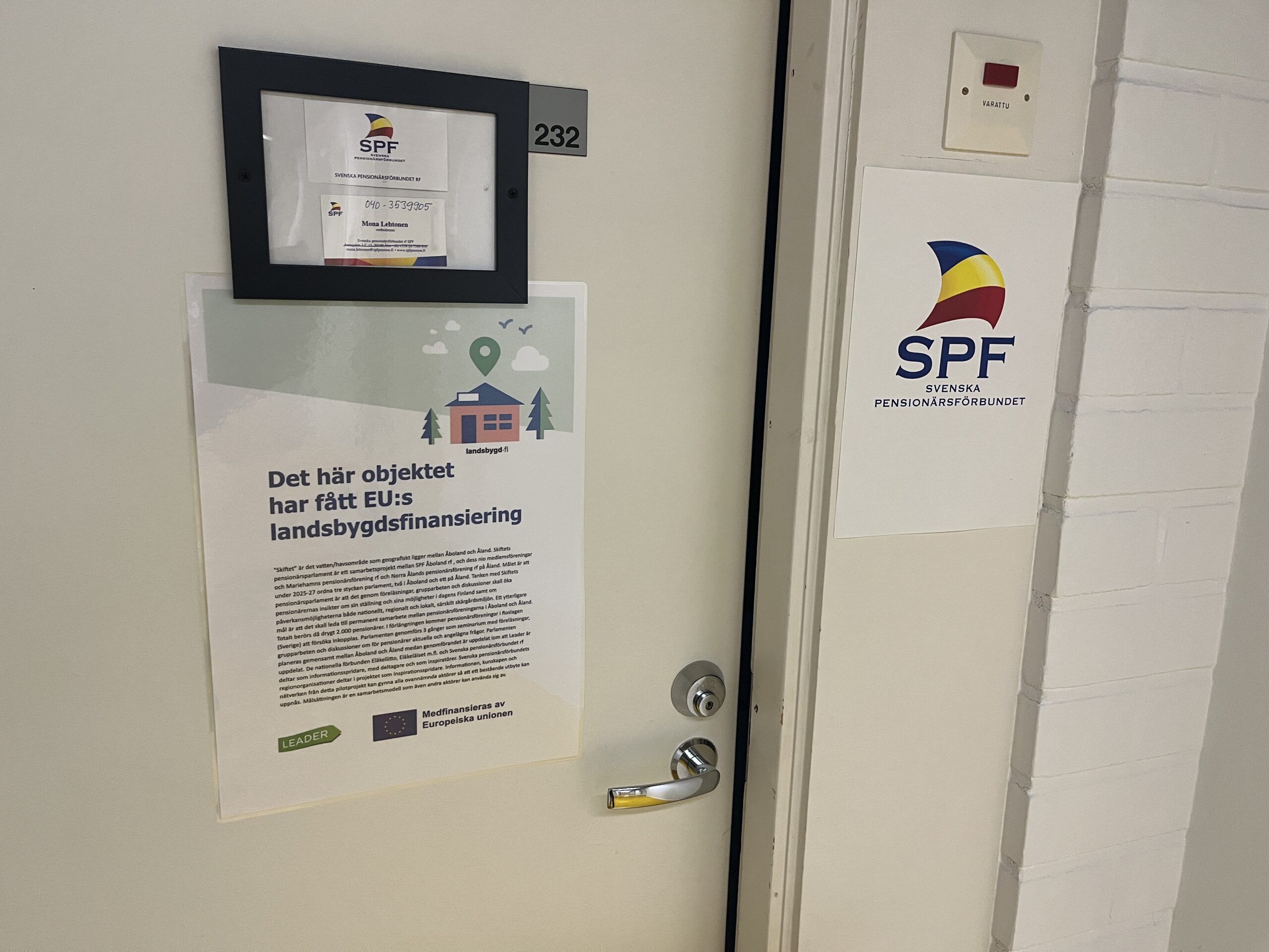 En vit dörr märkt med nummer 232, med en skylt med SPF:s logotyp och en affisch på svenska om EU:s stöd till landsbygdsutveckling. En knappsats eller kontrollpanel är monterad på väggen bredvid dörren.