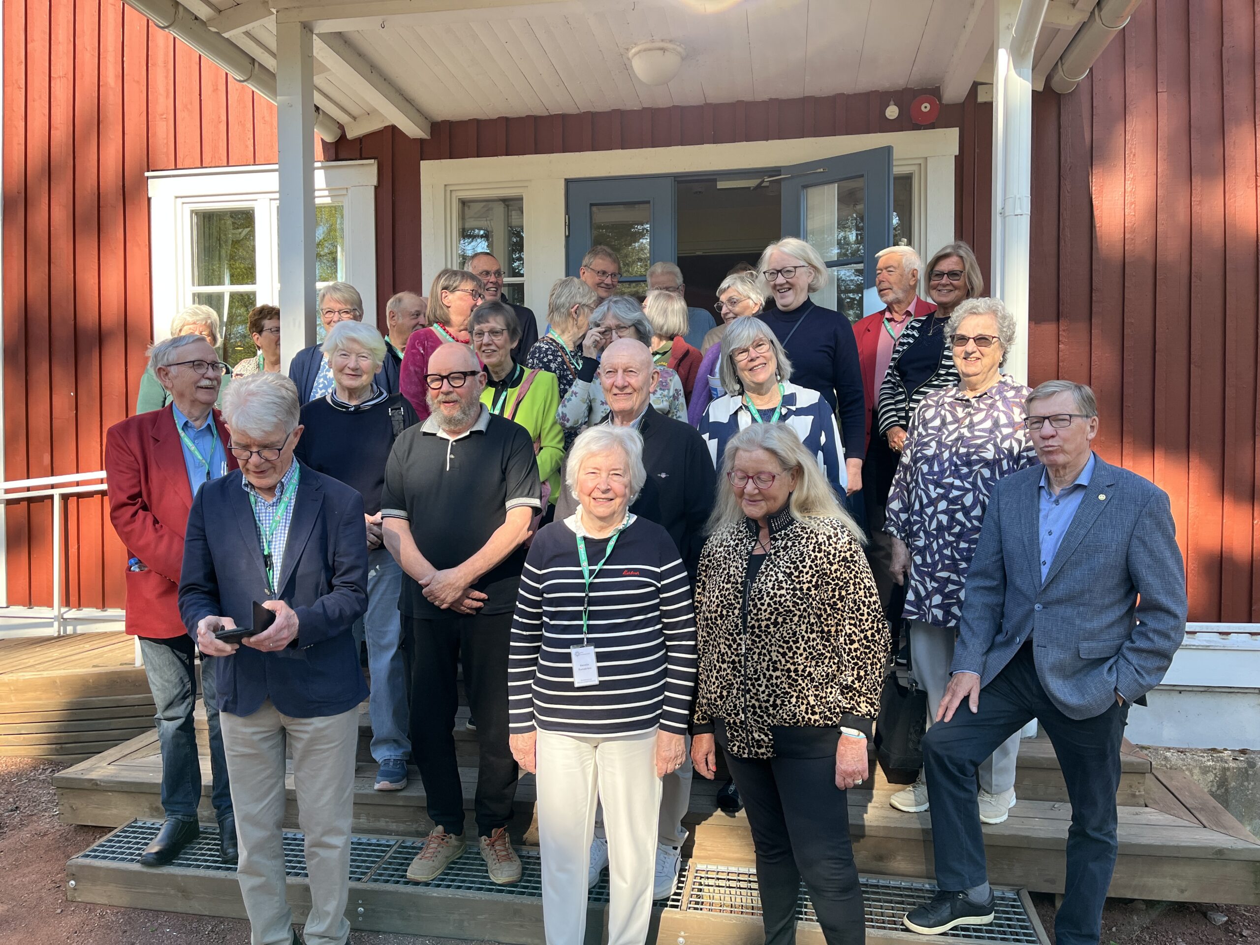 En grupp äldre vuxna står och sitter på trappan utanför en röd träbyggnad med vita kanter. De flesta ler och är vända mot kameran en solig dag.
