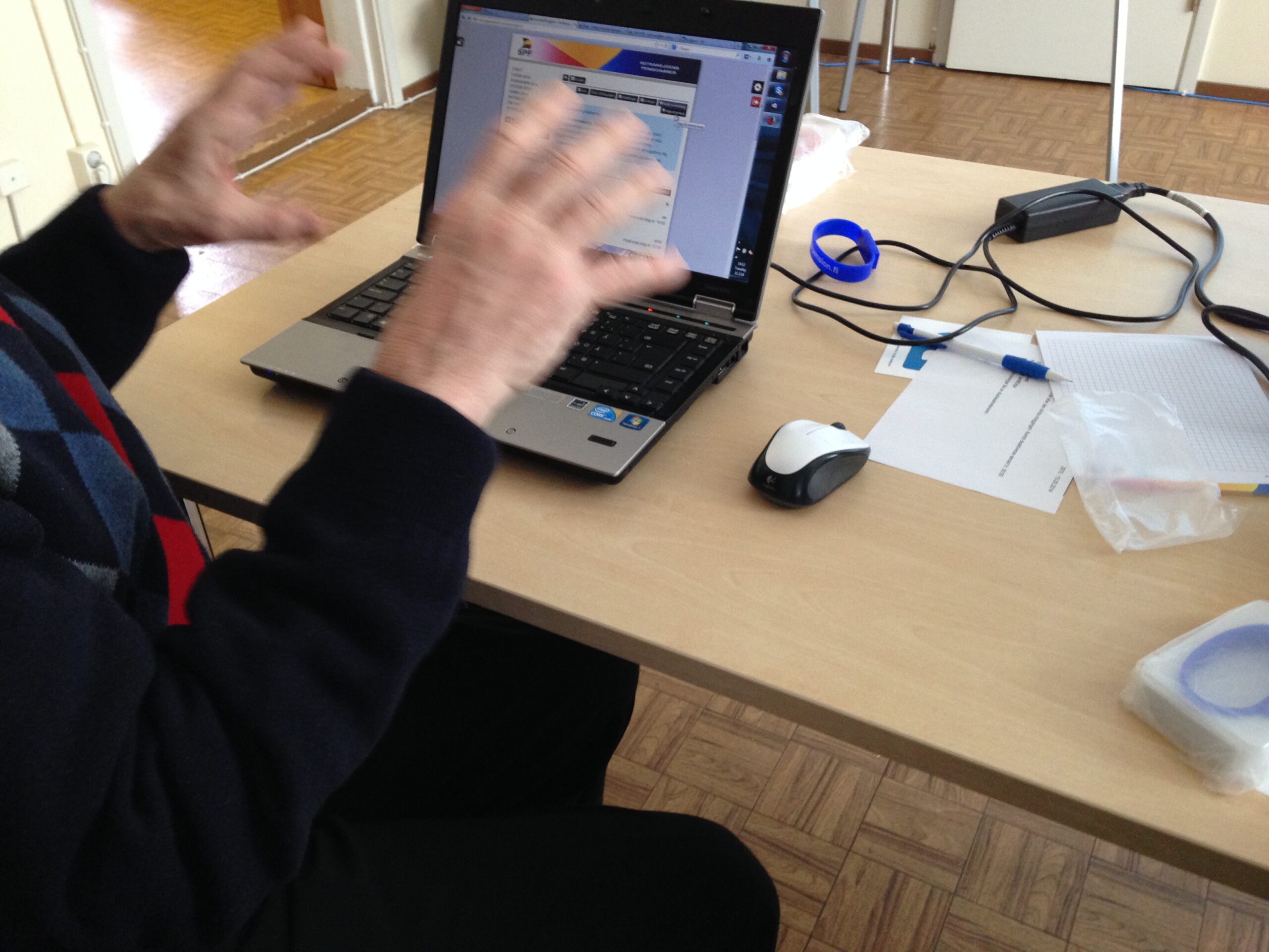 En person sitter vid ett skrivbord och använder en bärbar dator, med händerna suddiga i rörelse. På bordet ligger en datormus, ett anteckningsblock, en penna och några små verktyg. I bakgrunden syns ett trägolv och en del av en dörr.