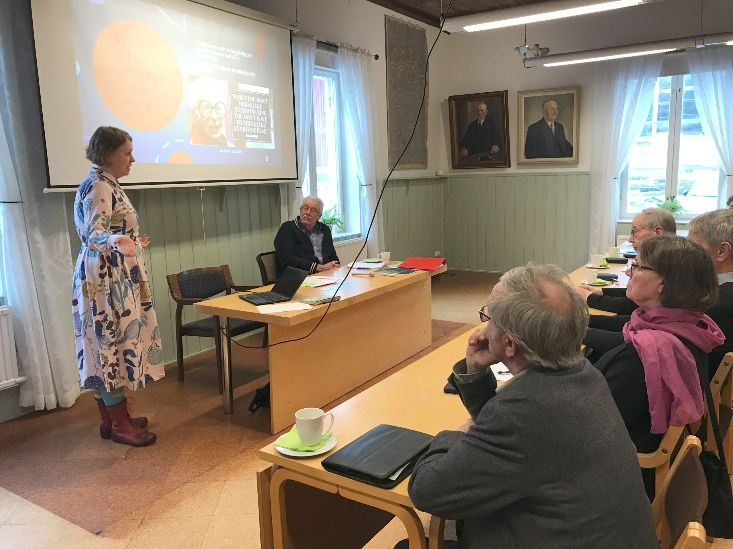 En kvinna står och talar till en grupp människor som sitter i ett klassrum eller konferensrum, med en presentation projicerad på en skärm bakom henne och flera porträtt upphängda på väggarna.