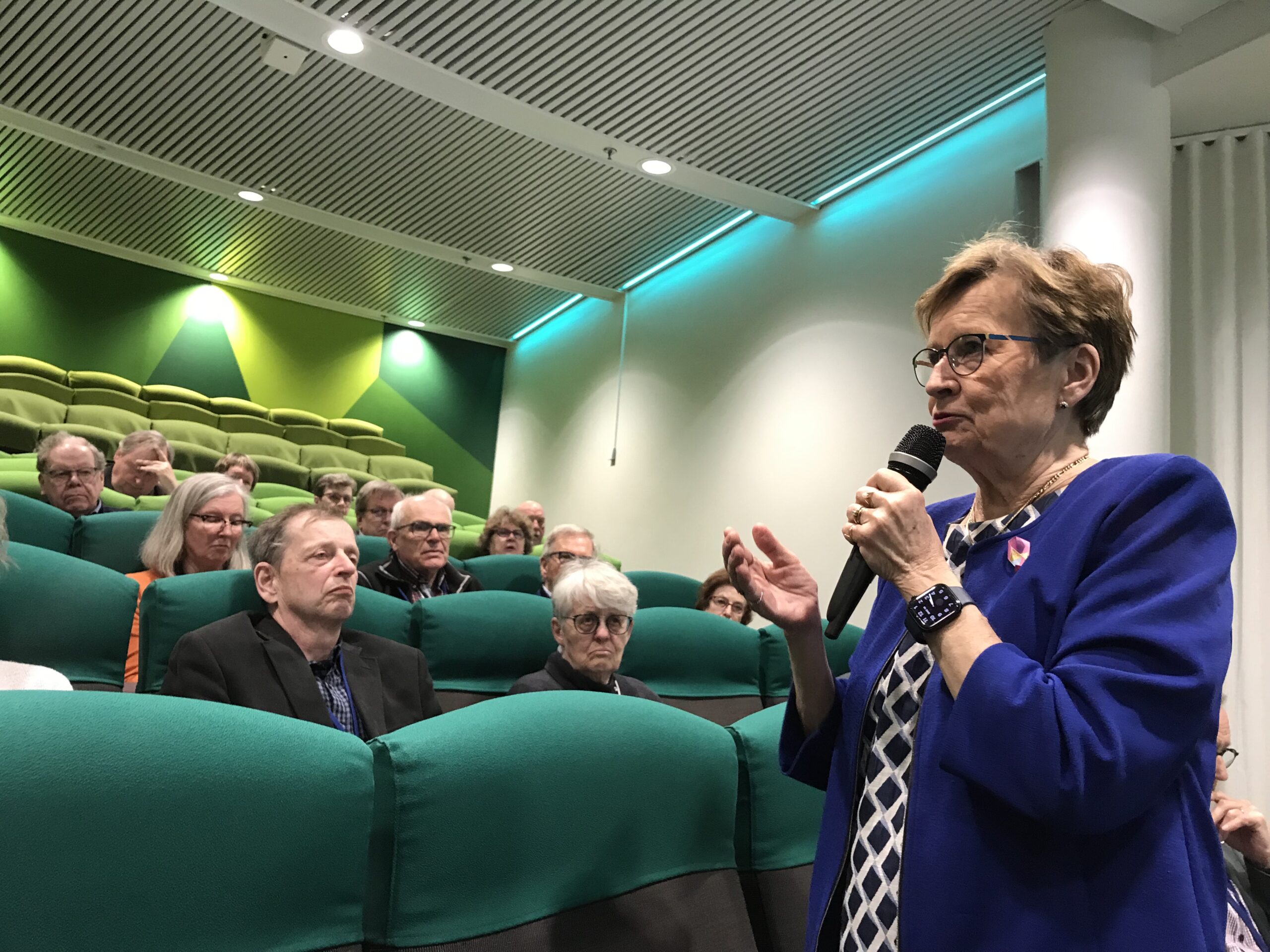 En äldre kvinna i en blå kavaj talar i en mikrofon medan hon står bland sittande människor i en sal med grön matta och modern belysning. Publiken lyssnar uppmärksamt.