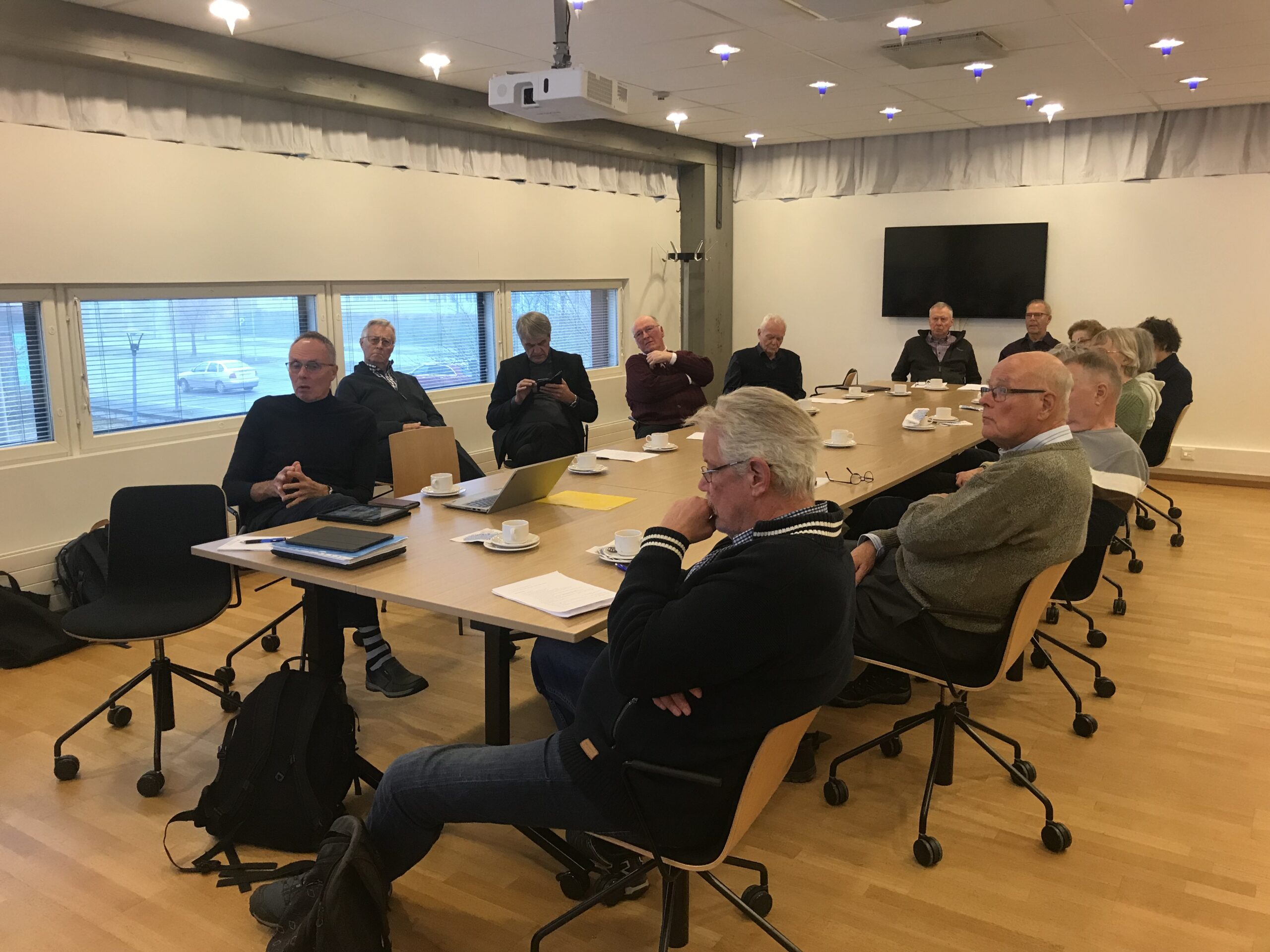 En grupp äldre vuxna sitter runt ett stort rektangulärt bord i ett mötesrum; några har bärbara datorer och papper framför sig. Det står kaffekoppar på bordet, och på väggen i bakgrunden hänger en stor skärm.