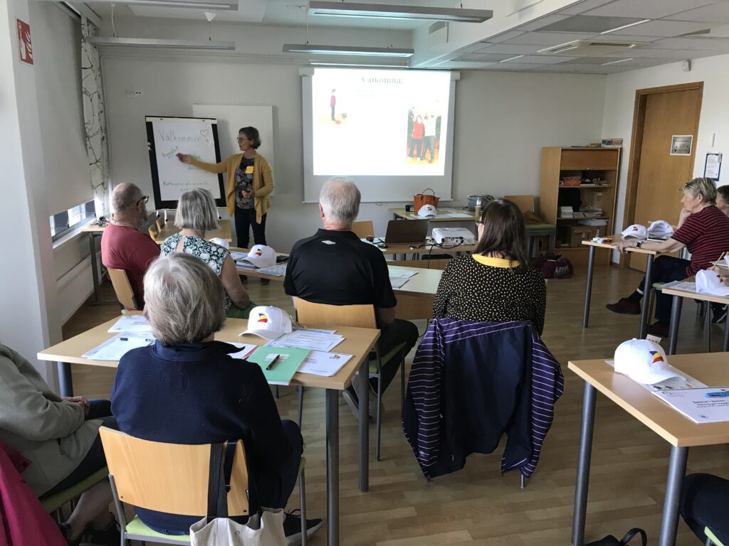 En kvinna står längst fram i ett klassrum och håller en presentation för en grupp sittande vuxna. På bänkarna ligger papper, mappar och vita mössor, medan en projektor visar bilder på en skärm bakom henne.