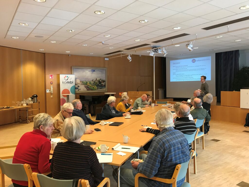 En grupp äldre vuxna sitter runt ett konferensbord och lyssnar på en föreläsare som står bredvid en projicerad bild. På bordet ligger anteckningsböcker, muggar och papper. Rummet är väl upplyst och har träpaneler på väggarna.
