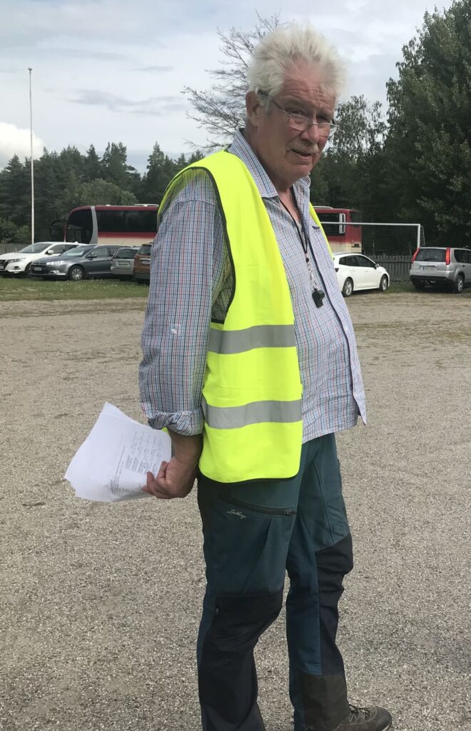 En äldre man med vitt hår och mustasch, som bär glasögon och en gul reflexväst och håller i några papper, står utomhus på en grusbelagd yta med parkerade bilar och en röd buss i bakgrunden.