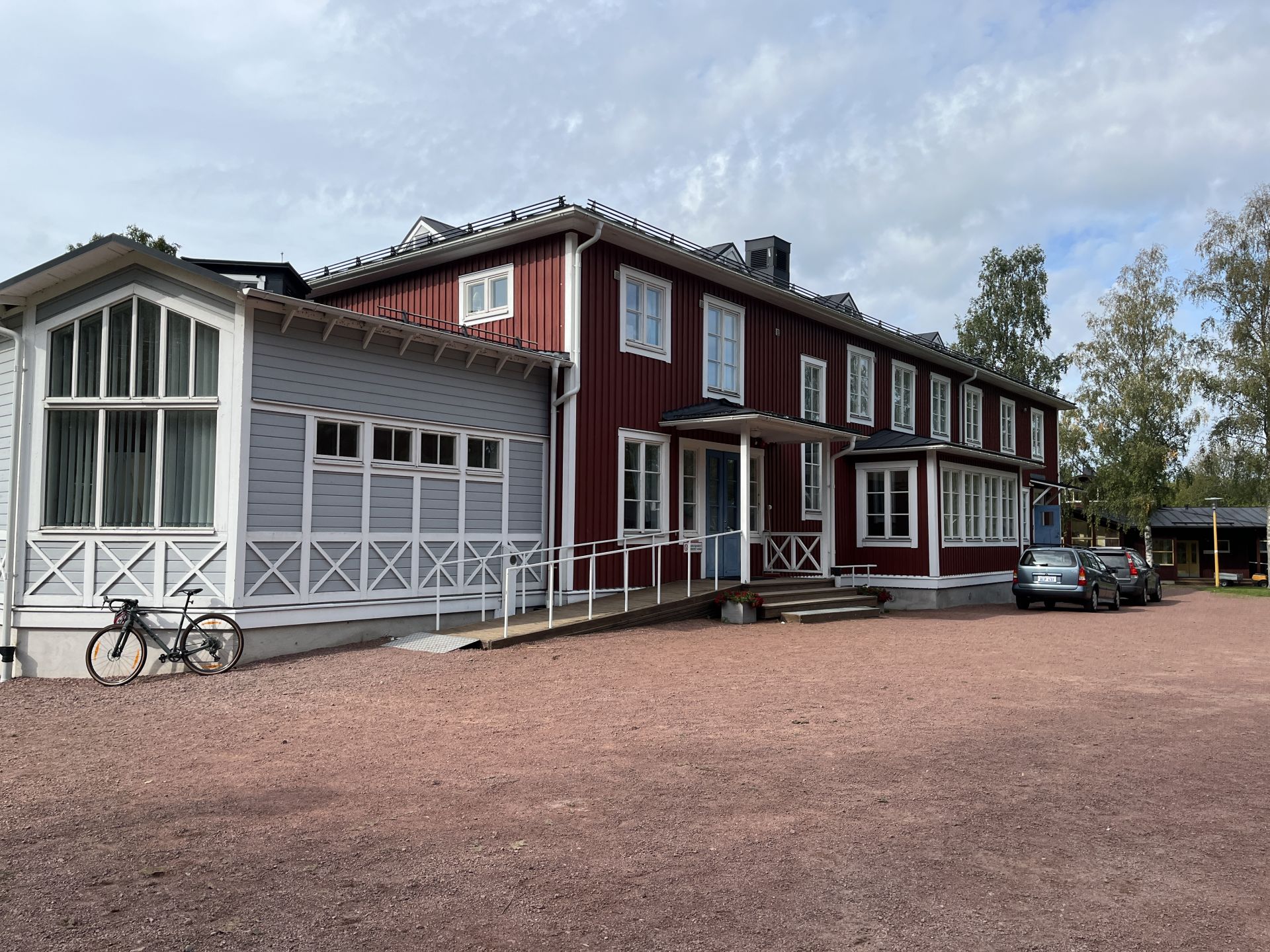 En tvåvåningsbyggnad i trä, målad i rött och vitt, med en ramp och trappsteg vid ingången, cyklar parkerade i närheten, två bilar i bakgrunden och träd som omger området under en delvis molnig himmel.