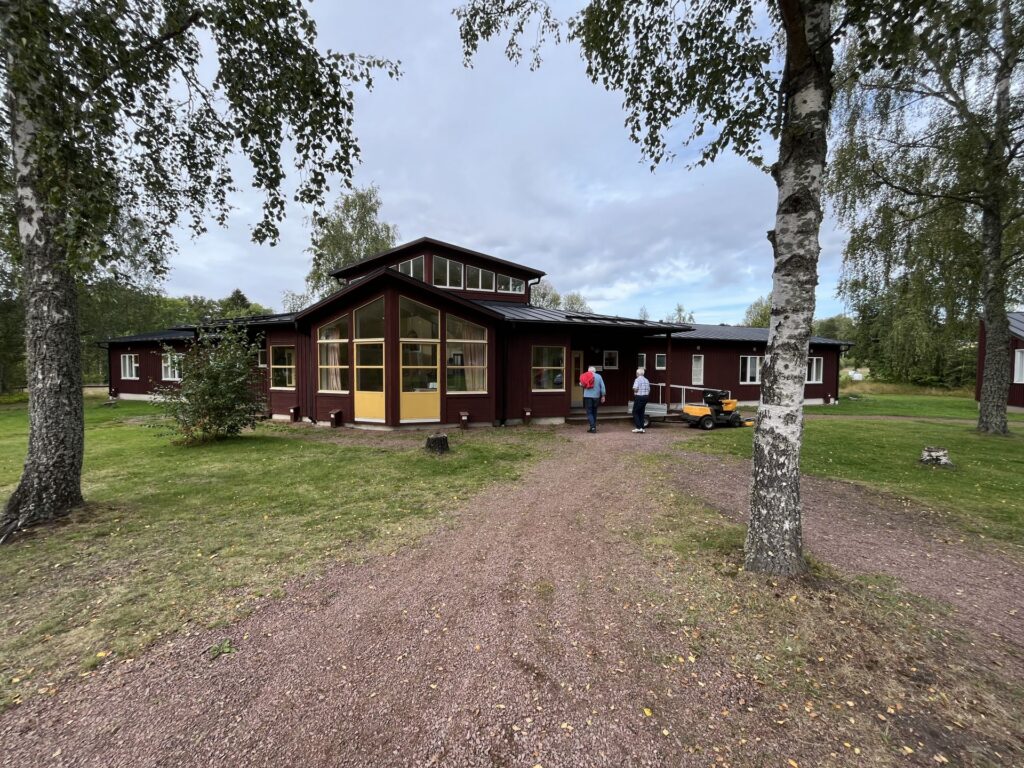 En brun träbyggnad med stora fönster och ett spetsigt tak står på en gräsbevuxen yta med träd. Två personer går längs en grusgång mot ingången, och en skottkärra syns på gräsmattan. Himlen är molnig.