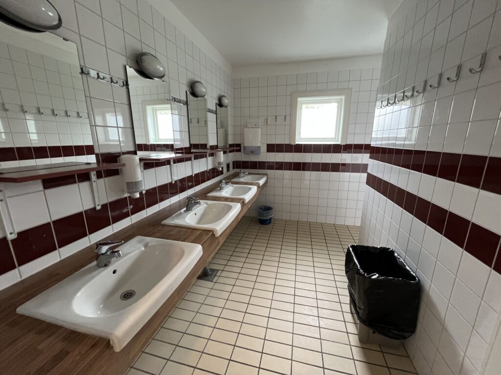 En offentlig toalett med tre vita handfat, speglar ovanför varje handfat, vita kaklade väggar med en röd rand, ett kaklat golv, ett fönster, tvåldispensrar, krokar på väggen, en papperskorg och en liten avfallskorg.