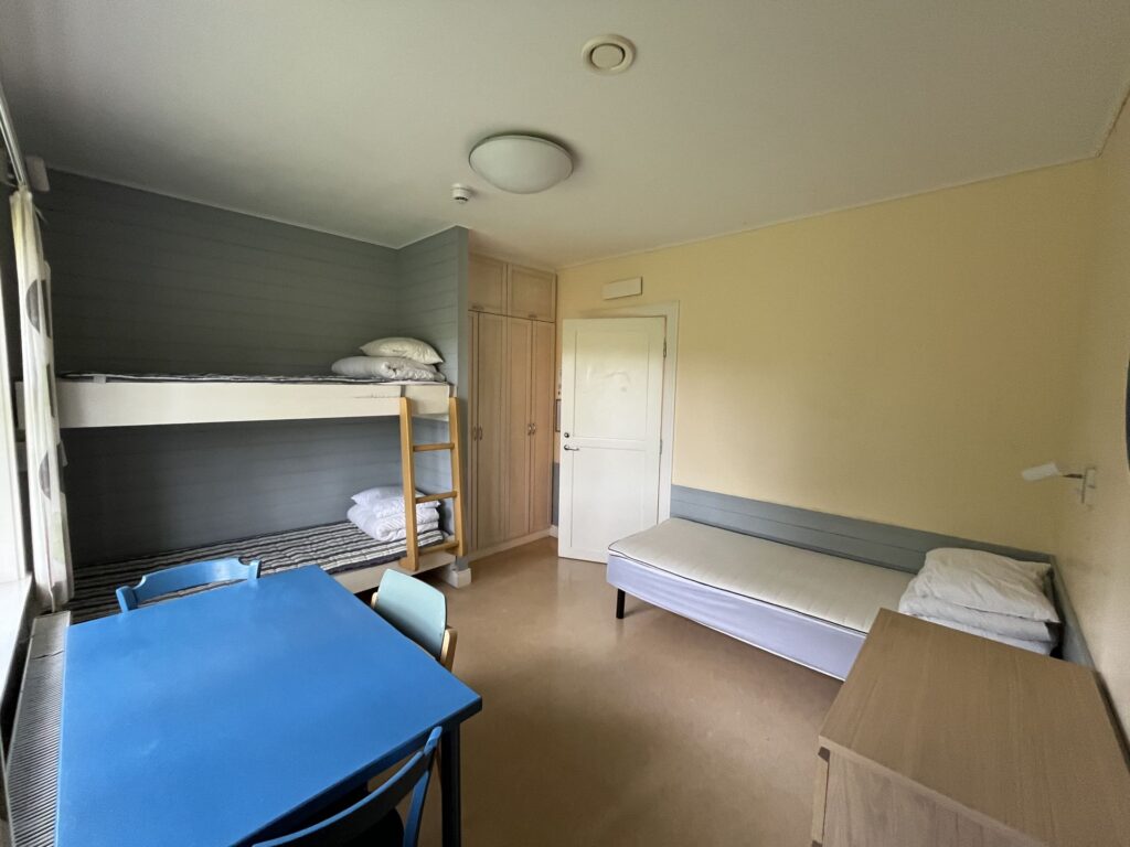 Ett litet, enkelt rum med en våningssäng, en enkelsäng, ett blått bord med fyra stolar, en byrå i trä och en garderob. Väggarna är ljusa och golvet är beige. Sängarna är bäddade med kuddar och vikta sängkläder.