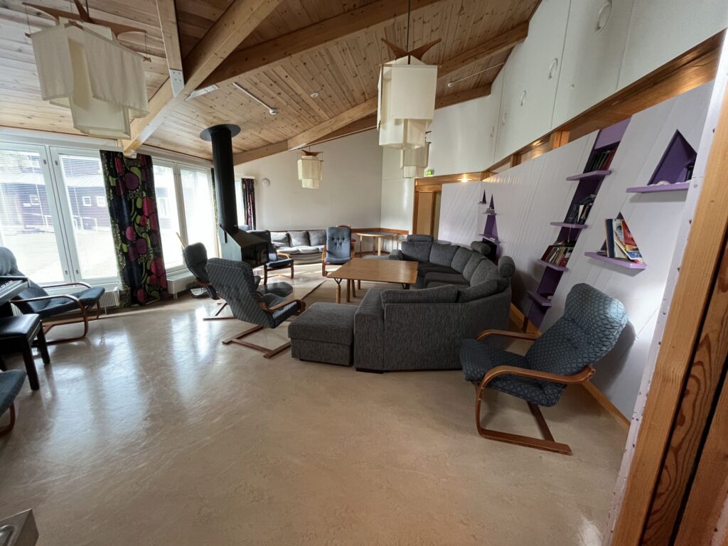 Ett rymligt vardagsrum med trätak, moderna lampor, en vedkamin, grå modulsoffor, fåtöljer, ett soffbord, bokhyllor med böcker och stora fönster med mönstrade gardiner.