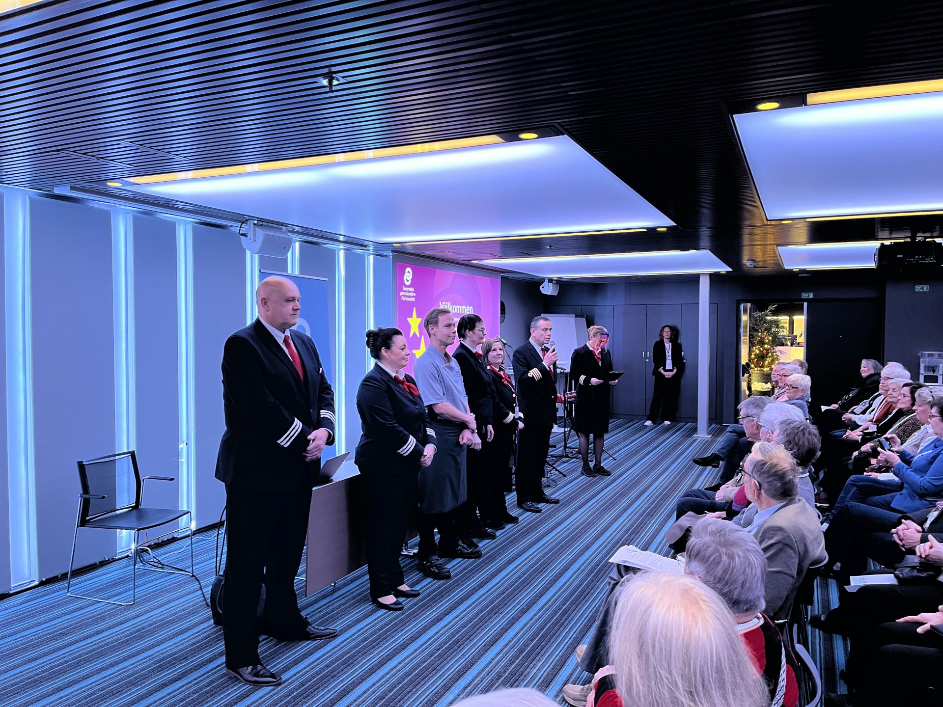 En grupp personer i uniform och formell klädsel står på rad inför en publik i ett modernt konferensrum med randig heltäckningsmatta, blå belysning och lila fond. Publiken lyssnar uppmärksamt.