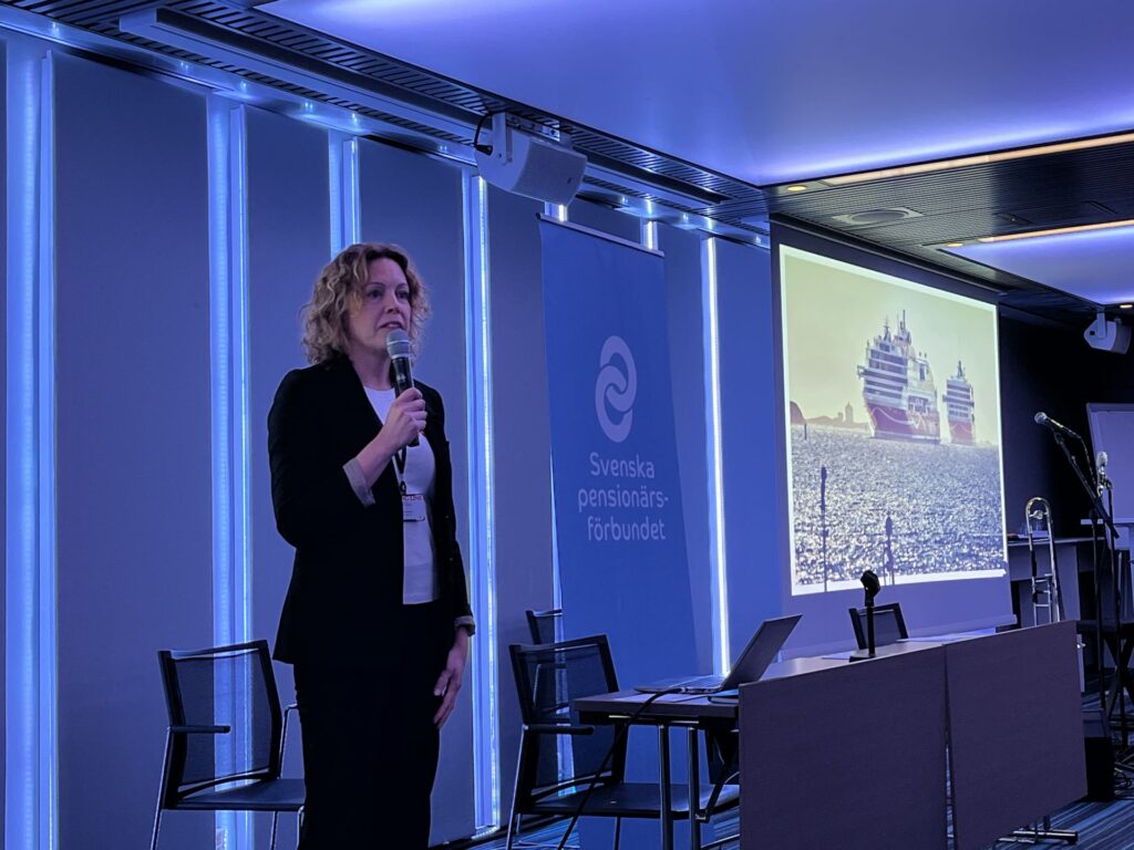 En person i affärskläder håller i en mikrofon och står nära ett podium i ett konferensrum med blå belysning. På en skärm visas en bild av kryssningsfartyg till havs. På en banderoll står det Svenska pensionärsförbundet.