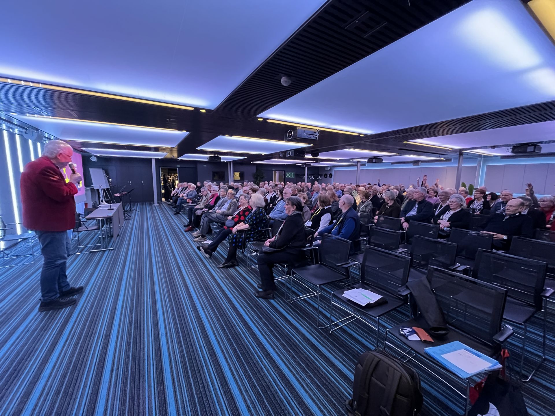 En man i röd kavaj talar till en sittande publik i ett modernt konferensrum med blårandig matta, takbelysning och presentationsutrustning längst fram. De flesta i publiken verkar vara äldre vuxna.
