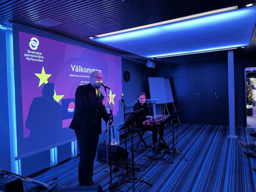En man i kostym talar i en mikrofon på en scen bredvid en keyboardist. Bakom dem finns en skärm där det står ”Välkommen” och Svenska pensionärsförbundets logotyp. Rummet är upplyst med blått ljus och där syns en julgran.