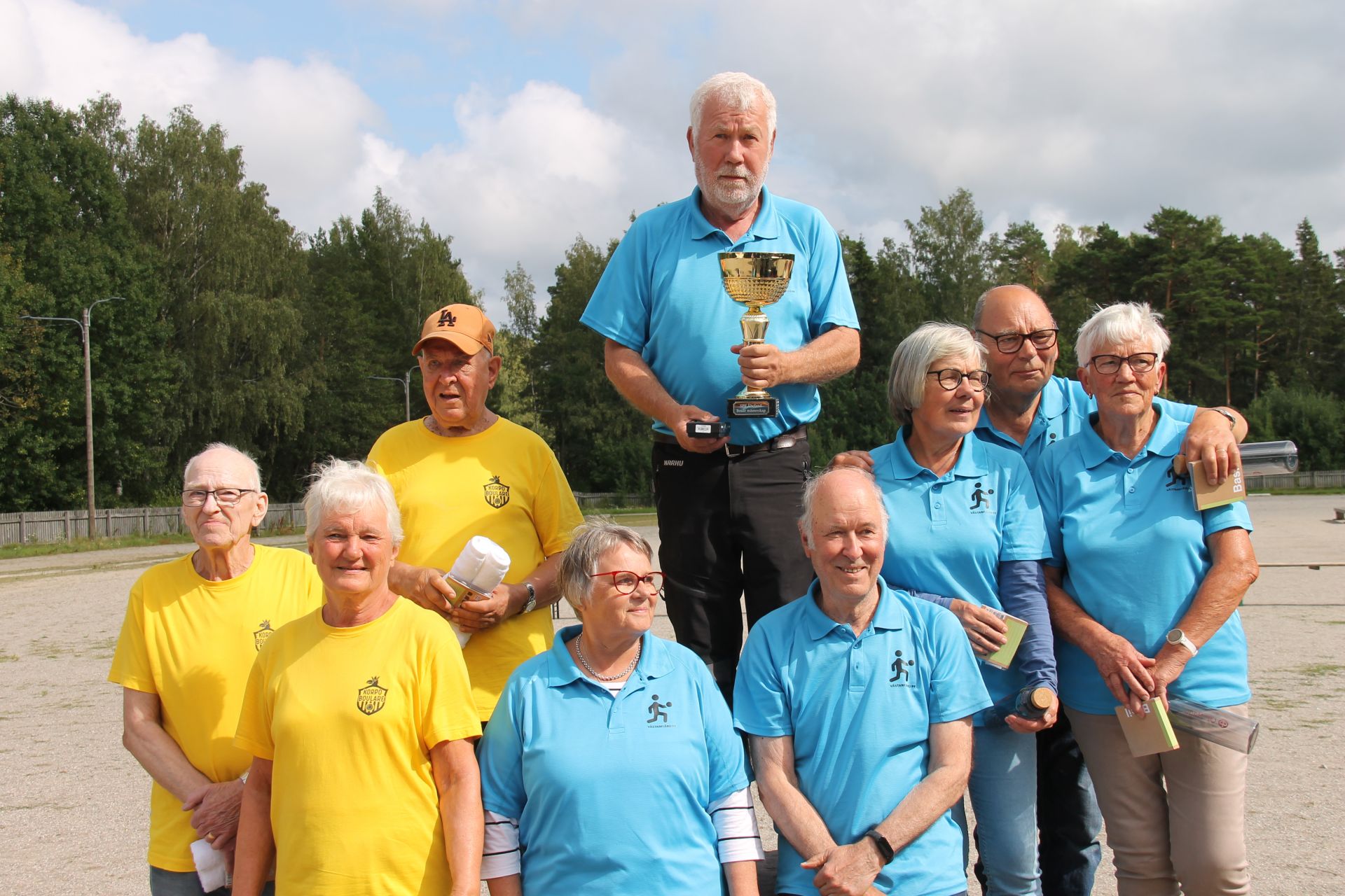 En grupp äldre vuxna poserar utomhus; fem av dem har blå tröjor och fyra har gula tröjor. En man i blå tröja står på en upphöjd plats i mitten och håller i en pokal. I bakgrunden syns träd och en molnig himmel.