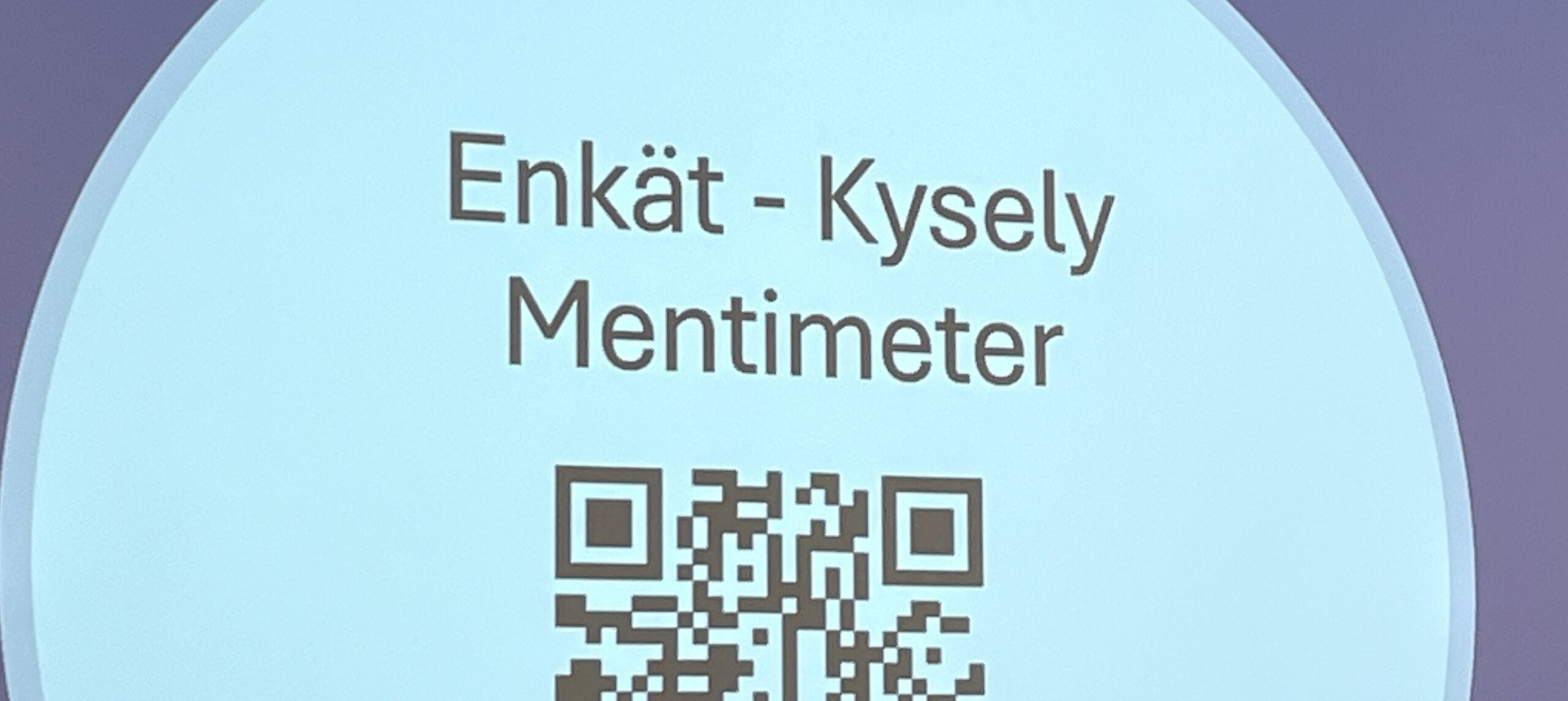 På skärmen visas texten ”Enkät – Kysely Mentimeter” ovanför en svartvit QR-kod, allt mot en ljusblå cirkulär bakgrund.