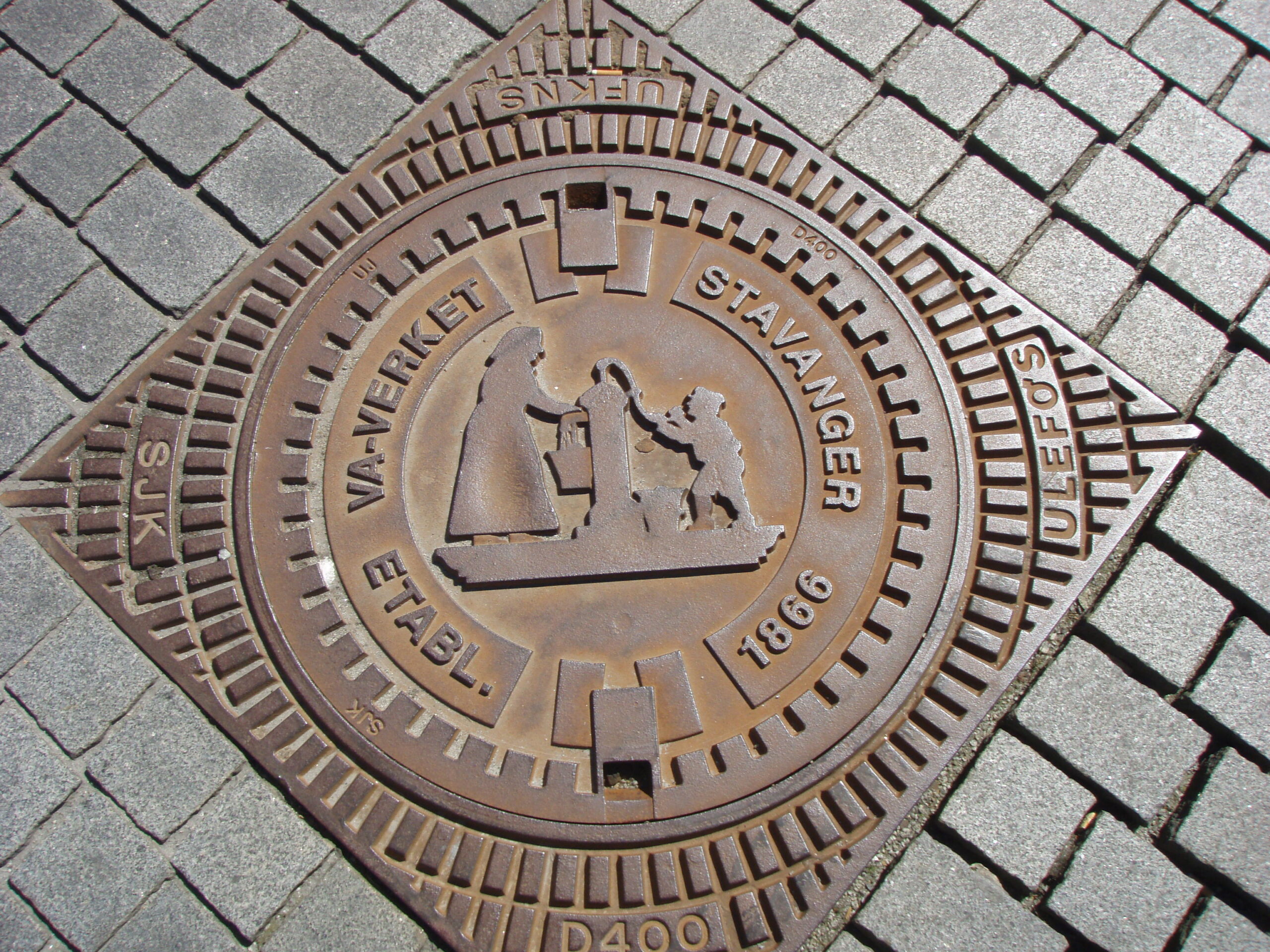 Rostfärgat brunnslock på en kullerstensbelagd trottoar. Locket är märkt VA-VERKET STAVANGER 1866 och har en upphöjd design av tre människofigurer samt ytterligare text längs kanten.