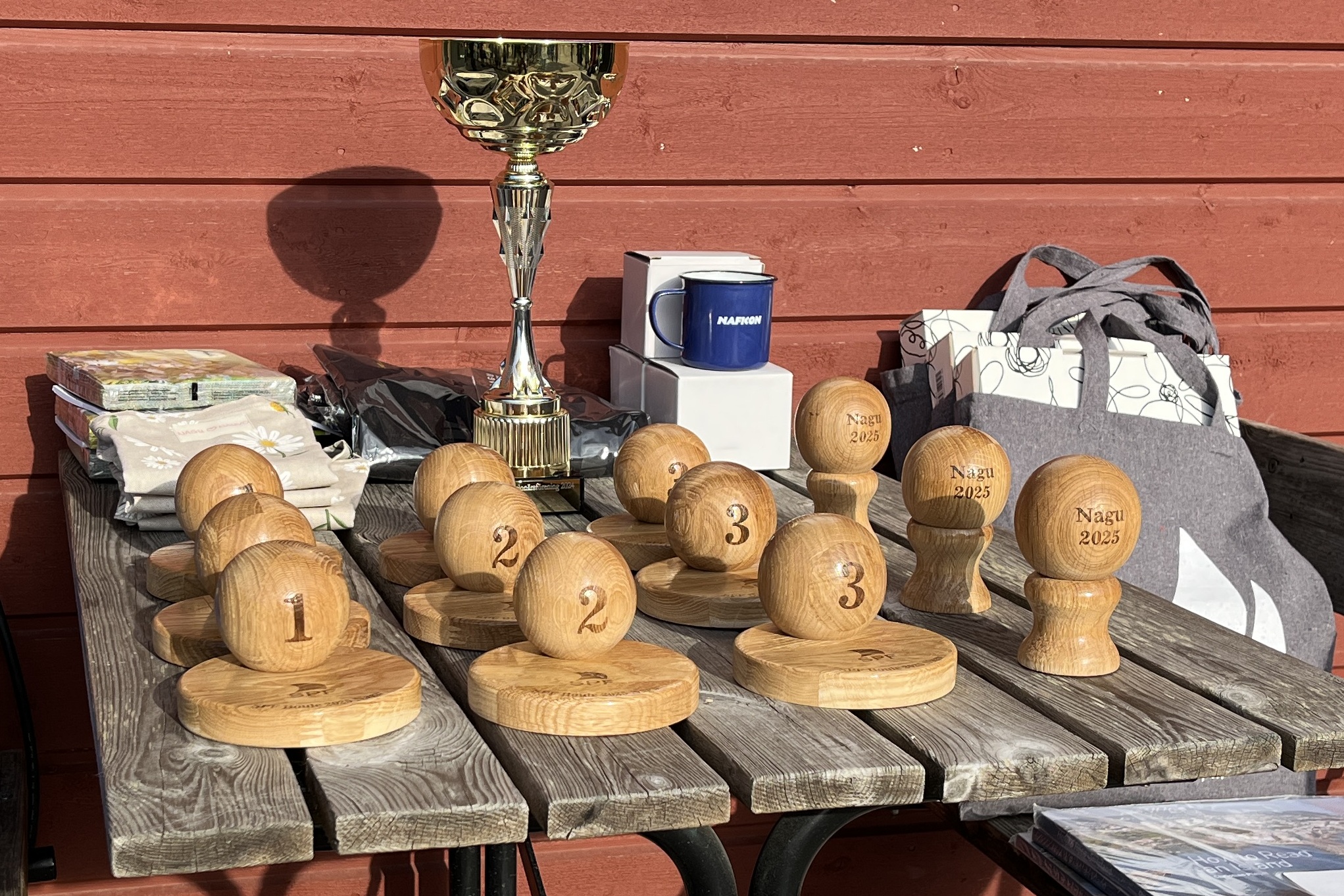 På ett träbord står flera trätroféer med ingraverade siffror och texten ”Nagu 2023”, en guldpokal, muggar, presentpåsar och en röd trävägg i bakgrunden. Solljuset kastar tydliga skuggor.