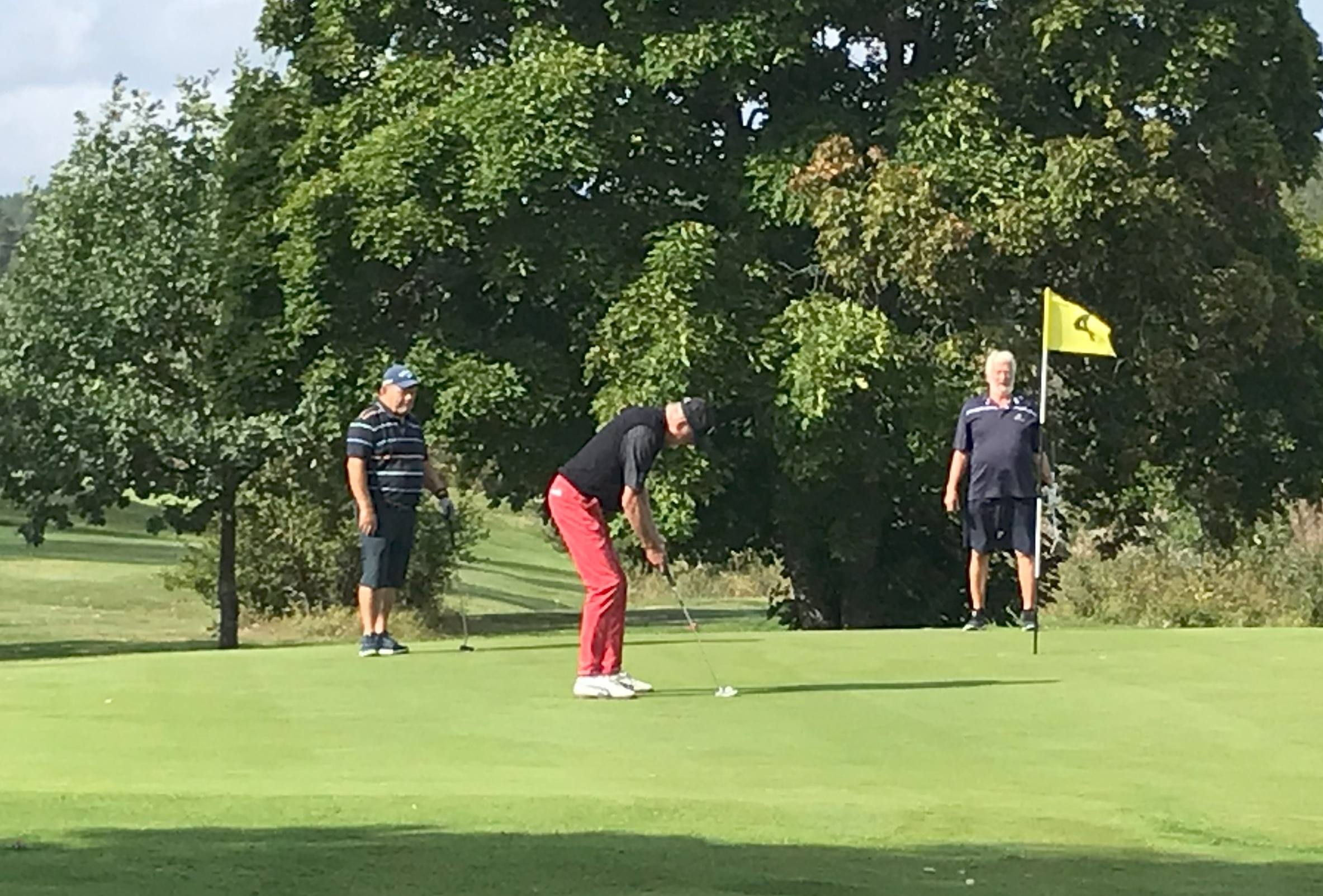 Tre personer på en golfbanas green; en person puttar medan två andra står i närheten och tittar på. En gul flagga markerar hålet, och i bakgrunden finns träd och gräs, under en delvis solig himmel.