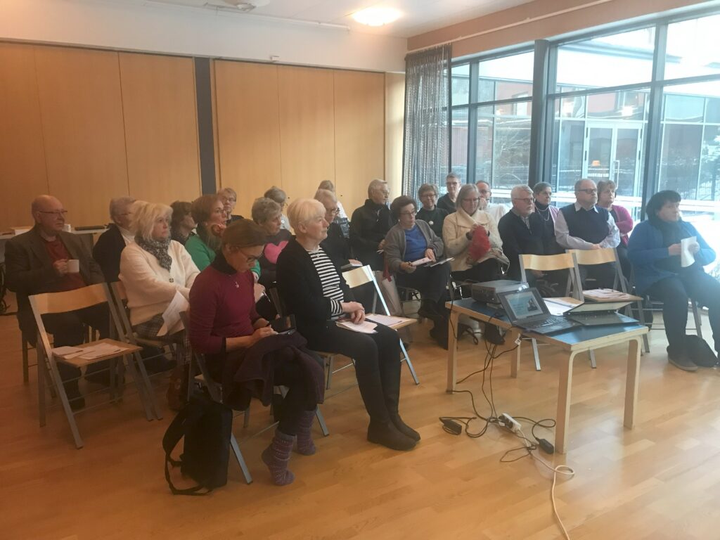 En grupp vuxna sitter på stolar i ett rum med trägolv och stora fönster, vända mot ett bord där det står en bärbar dator och en projektor. Vissa håller papper i händerna, och en strömkabel löper längs golvet.