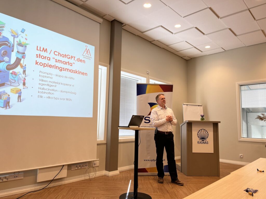 En man står framför en presentationsskärm i ett konferensrum. Bilden på skärmen har rubriken ”LLM / ChatGPT, den stora smarta kopieringsmaskinen” och innehåller punkter på svenska.