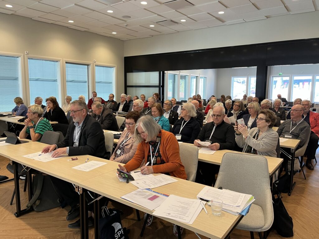 En grupp människor sitter vid långa bord i ett konferensrum och deltar i ett möte eller seminarium. Vissa antecknar, andra tittar på dokument eller elektroniska enheter. På borden ligger papper och vattenglas.