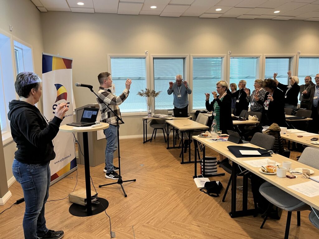 En grupp människor i ett konferensrum står upp och räcker upp händerna medan en person vid talarstolen leder en övning. Man ser bord med papper, tallrikar och drycker, och till vänster står en banderoll.