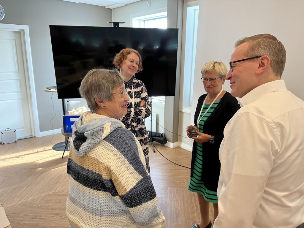 Fyra vuxna står i en liten grupp och samtalar i ett väl upplyst rum med en stor TV-skärm och fönster i bakgrunden. Två kvinnor vänder sig mot kameran, medan en man och en kvinna står delvis med ryggen vänd mot kameran.