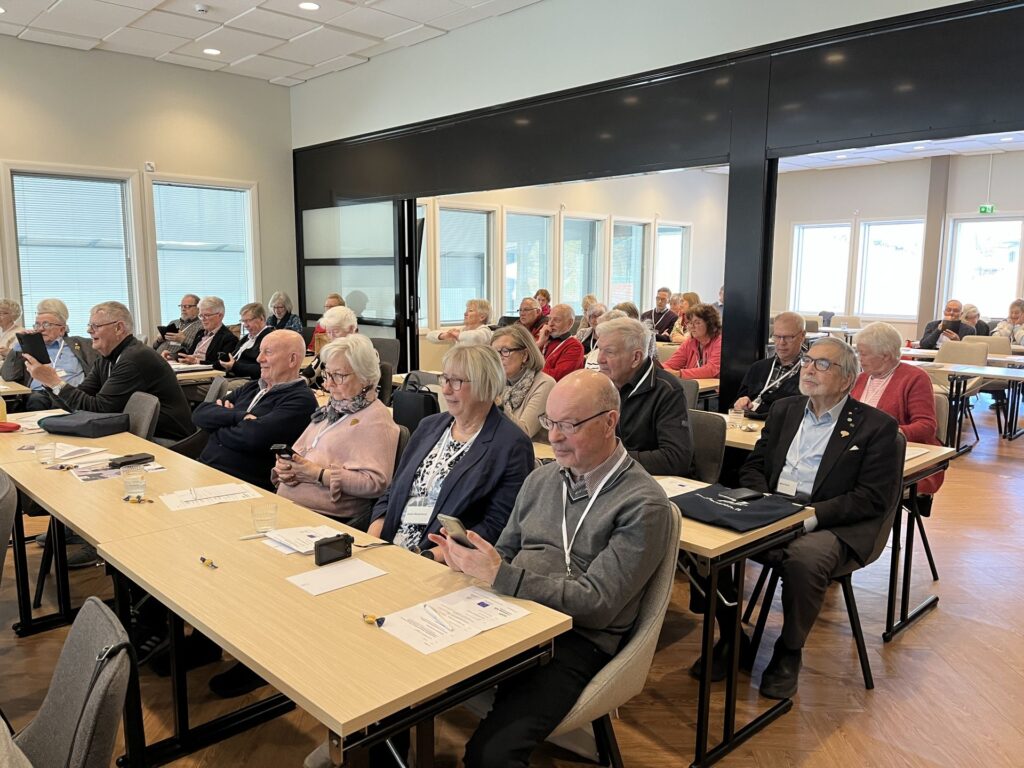 En grupp äldre vuxna sitter vid borden i ett konferensrum, vända framåt. Vissa tittar på sina mobiler eller anteckningar, medan andra följer med uppmärksamt. Rummet har stora fönster och väggar i neutrala färger.