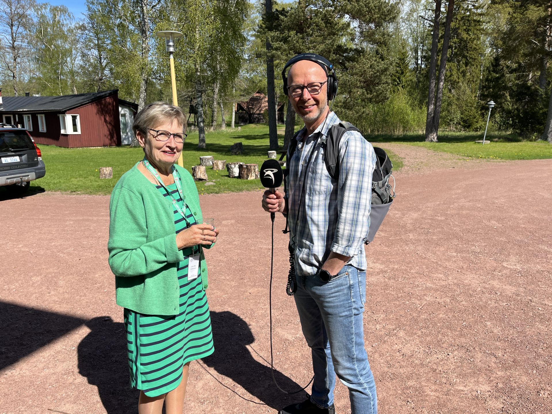 En kvinna i grön klänning och kofta står utomhus tillsammans med en man som bär hörlurar, glasögon, en rutig skjorta, jeans och ryggsäck. Mannen håller i en mikrofon. I bakgrunden syns träd och byggnader.