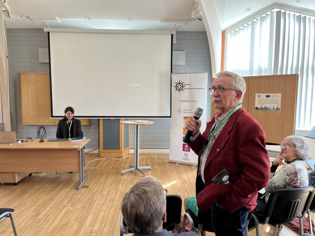 En äldre man i en röd jacka står med en mikrofon och talar till en sittande publik. En kvinna sitter vid ett skrivbord framför en tom projektorskärm. Övriga personer sitter och lyssnar i ett ljust, modernt rum.