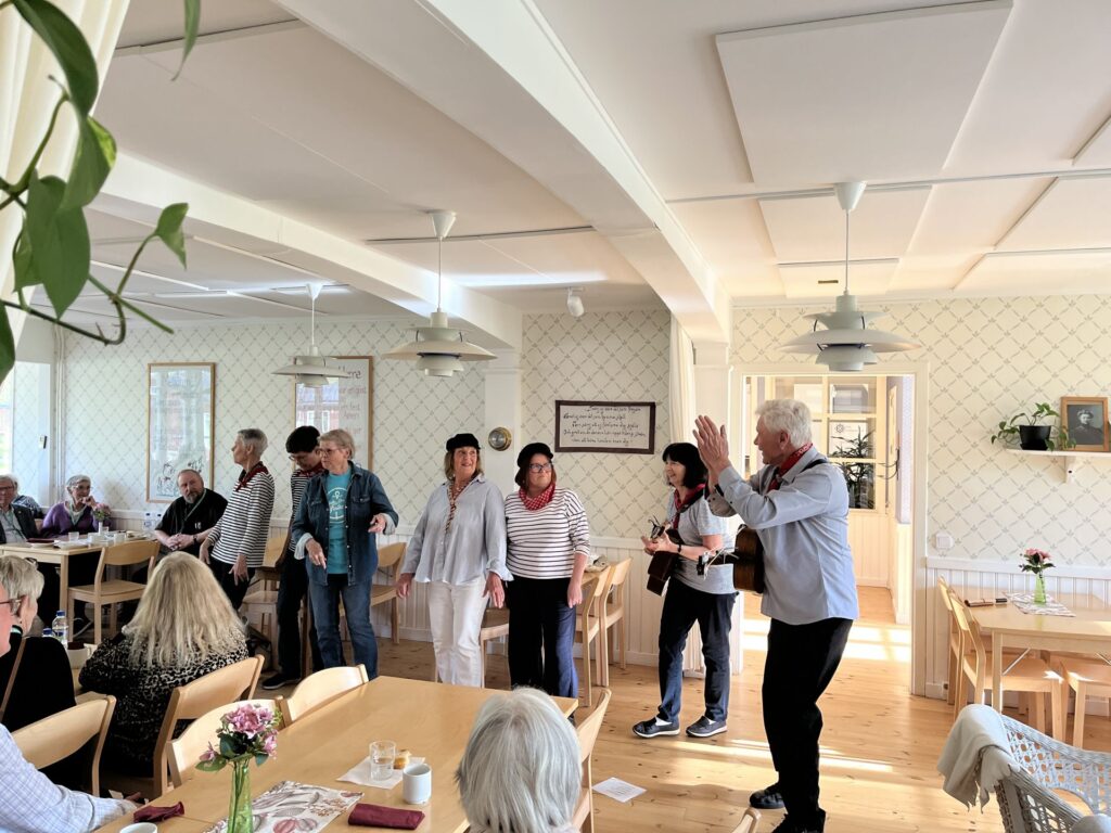 En grupp människor står och applåderar framför en grupp sittande personer i ett ljust, mysigt rum med trägolv och bord. Vissa pratar, medan andra tittar på. På borden står blommor i vaser.
