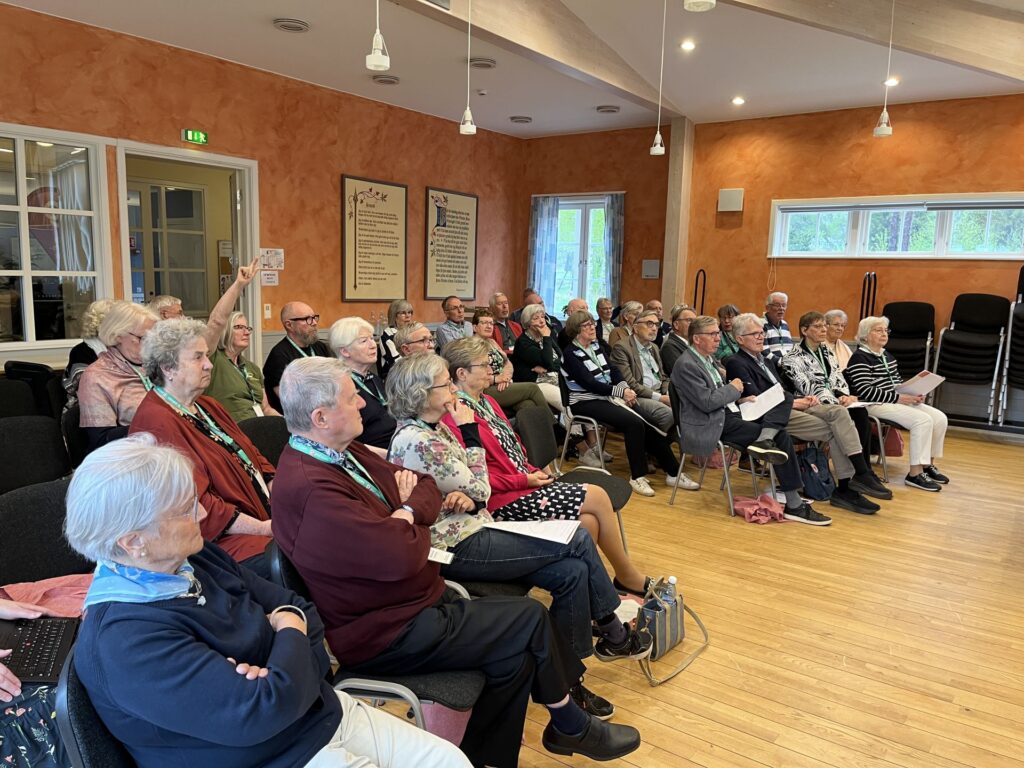 En grupp äldre sitter i rader av stolar i ett ljust rum med orange väggar och lyssnar uppmärksamt. Vissa håller i papper eller anteckningsböcker, och några bär gröna nyckelband. I hörnet står staplade tomma stolar.