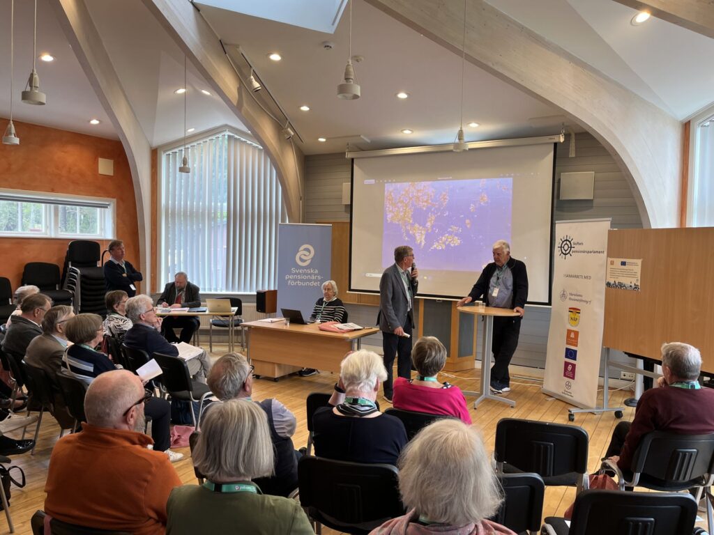 En grupp vuxna som sitter i ett konferensrum lyssnar på två män som talar längst fram, med presentationsbanderoller och en stor skärm som visar en karta bakom dem. Rummet har välvda träbjälkar och stora fönster.