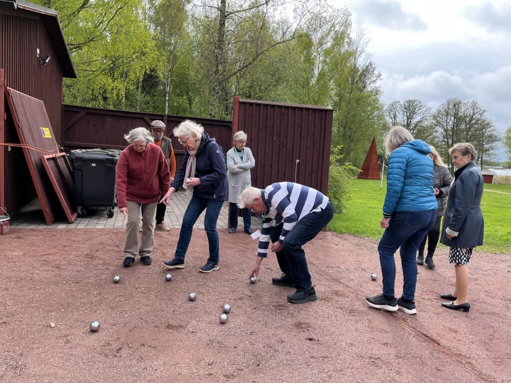 En grupp äldre står utomhus på en grusplan, böjer sig ner och tittar på metallkulor på marken; det ser ut som om de spelar pétanque. I bakgrunden syns träd och träbyggnader.