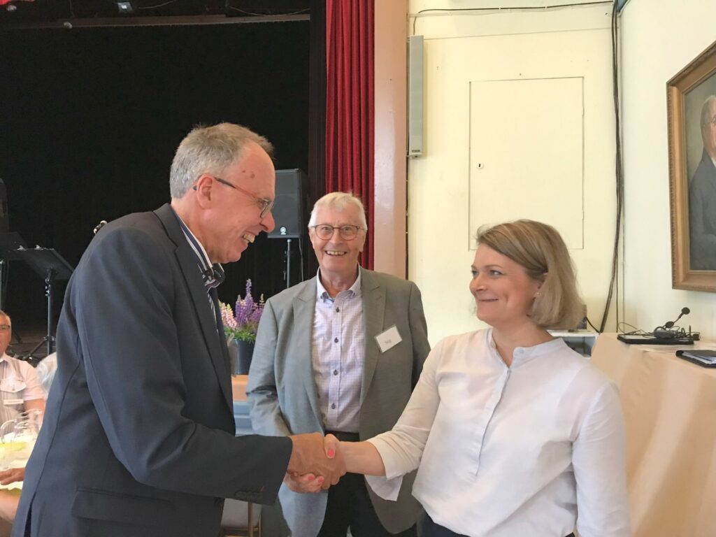 En man i kostym skakar hand med en kvinna i vit blus vid ett evenemang inomhus. En annan man i grå kavaj står mellan dem och ler. I bakgrunden syns människor, bord och ett porträtt.