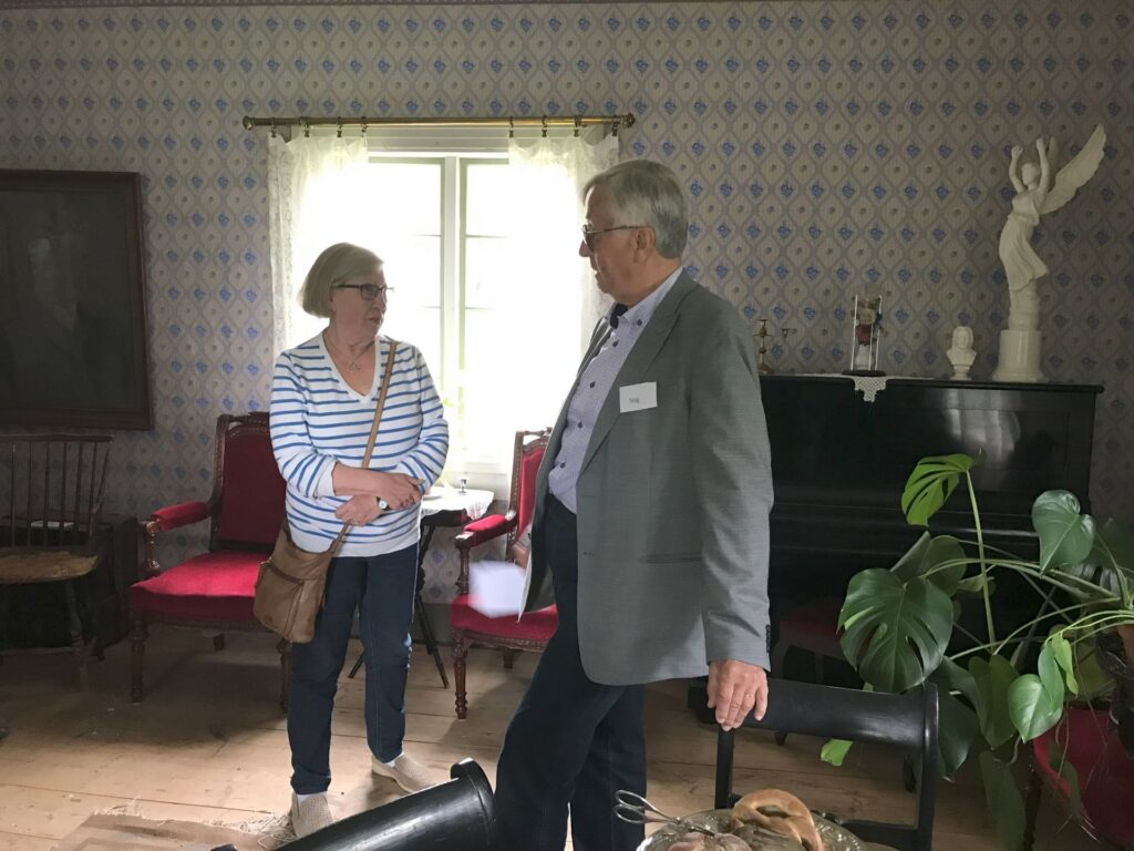 En kvinna och en man står och pratar i ett rum med mönstrad tapet, ett fönster med tunna gardiner, röda stolar, ett piano, en krukväxt och en vit staty ovanpå pianot.