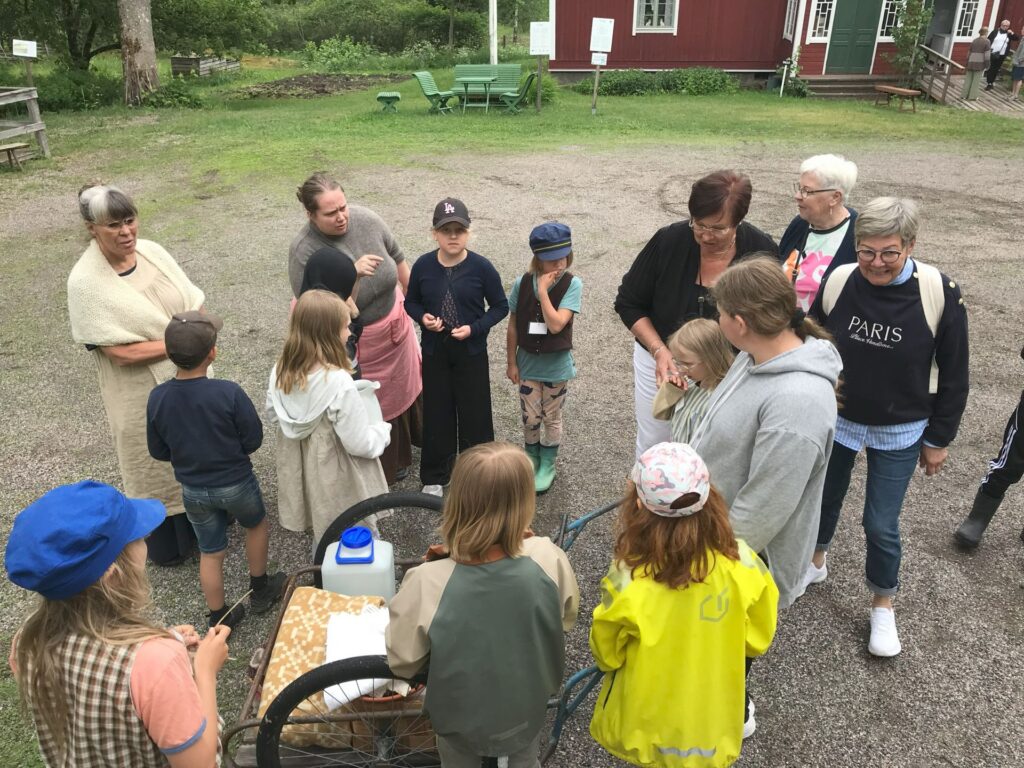 En grupp vuxna och barn har samlats utomhus på en grusplan nära gröna stolar och röda byggnader. Vissa barn har på sig mössor och jackor, och i förgrunden står en vagn med en vattenbehållare.