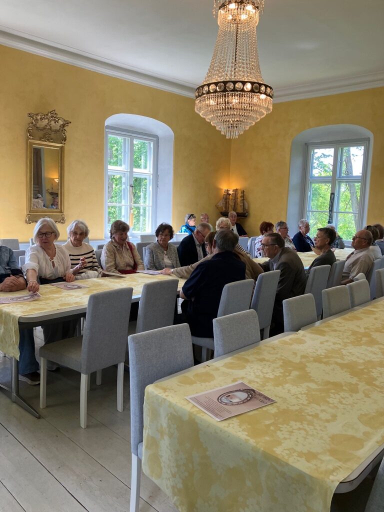En grupp äldre personer sitter runt ett långbord i ett rum med gula väggar, stora fönster och en kristallkrona. Några pratar medan andra tittar framåt. Bordet är täckt med en gulmönstrad duk.