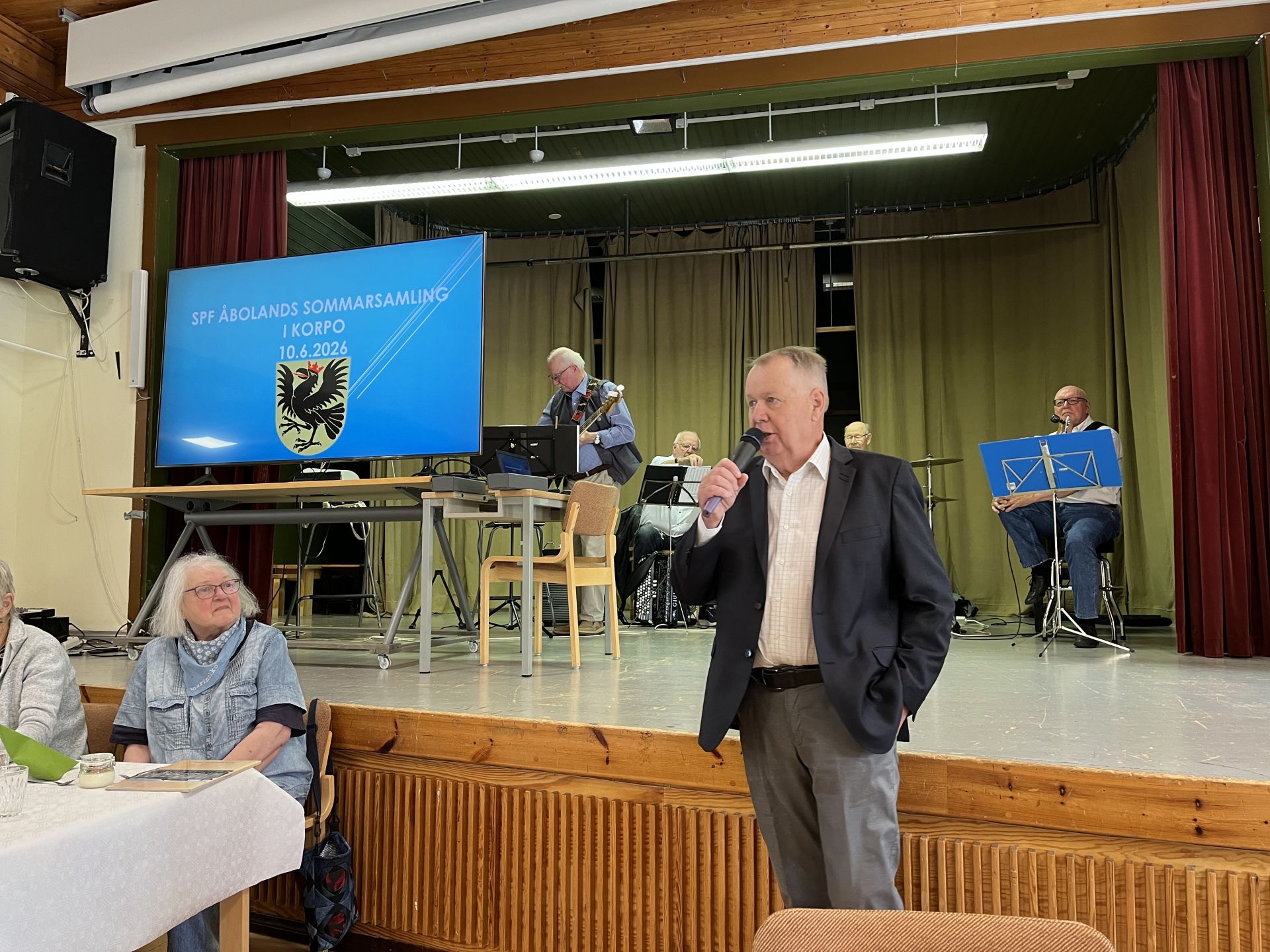 En kostymklädd man talar i en mikrofon i en sal. Bakom honom spelar musiker instrument på en scen med en stor skärm som visar SFF Ålands sommarsamling i Kökar 10.6.2026 och ett örnemblem. Människor sitter vid bord i förgrunden.