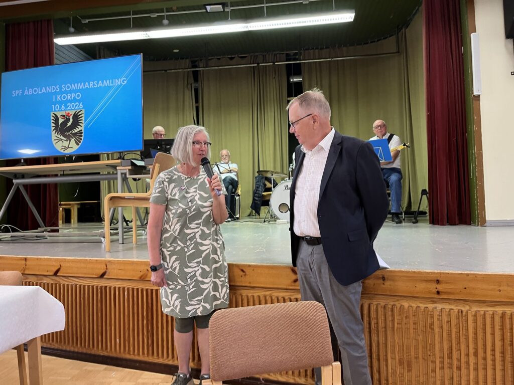 En kvinna med mikrofon talar till en kostymklädd man på en scen. Bakom dem sitter tre musiker med instrument. På en storbildsskärm visas texten "SFP Åbolands Sommarssamling i Korpo 10.6.2026.".